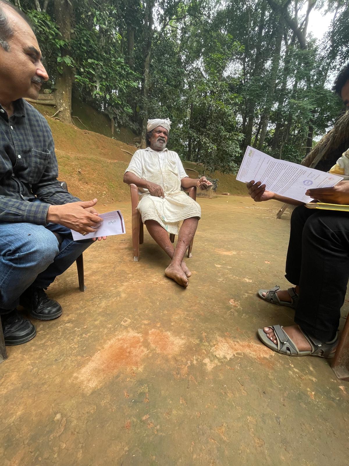 Cheruvayal Raman seated outdoors talking with two people holding survey documents
